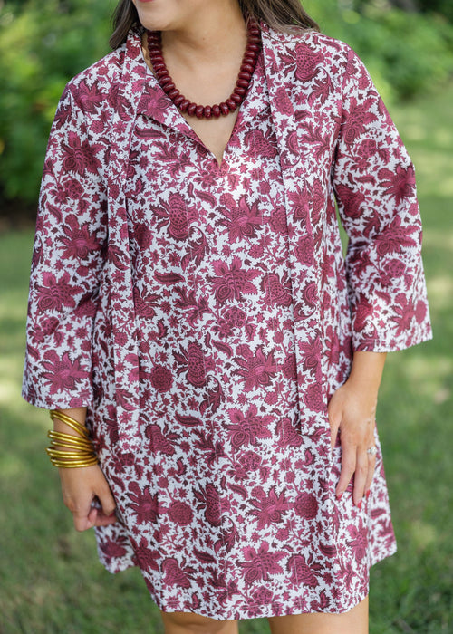 A woman wearing a short-sleeve collared tunic dress with a pink and white floral pattern, standing outdoors on a grassy lawn.