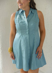 A woman wearing a sleeveless, button-front mini dress with vertical blue and white pinstripes, standing against a textured off-white wall.
