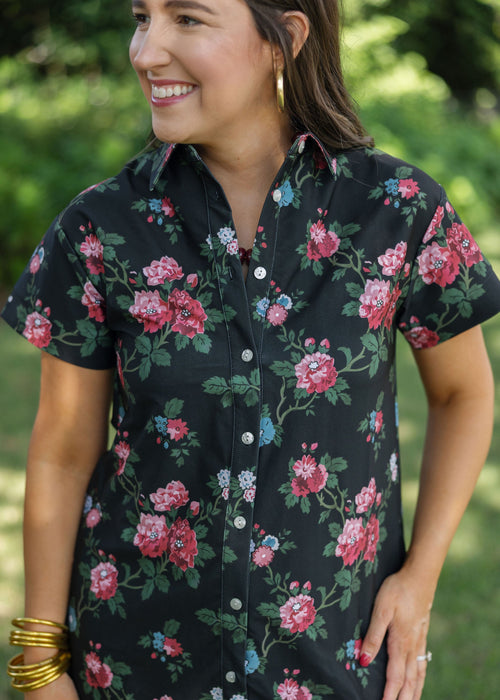 A woman wearing a short-sleeve button-up shirt dress with a black base and a vibrant pink, red, and blue floral print, photographed outdoors against a blurred green background.
