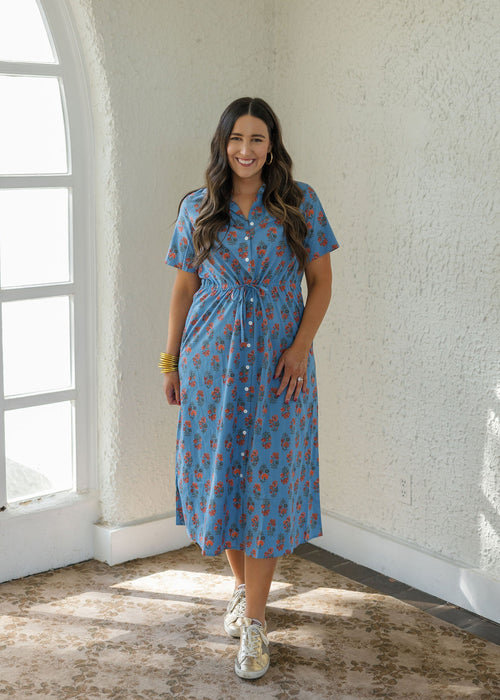 A woman wearing a blue short-sleeve button-up midi dress with a red and yellow pattern, standing in a bright room with textured white walls and a large arched window.