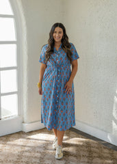 A woman wearing a blue short-sleeve button-up midi dress with a red and yellow pattern, standing in a bright room with textured white walls and a large arched window.
