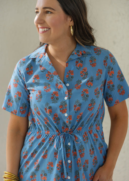 A woman wearing a short-sleeve button-front dress with a blue base and orange floral print, standing against a light neutral wall.