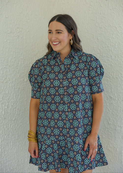 A woman wearing a short-sleeve button-up dress with a blue, teal, and white geometric floral pattern, standing against a textured off-white wall.