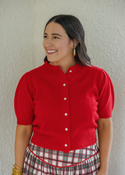 A woman wearing a bright red short-sleeve knit cardigan with white buttons and a red-and-white plaid skirt, standing against a textured off-white wall.