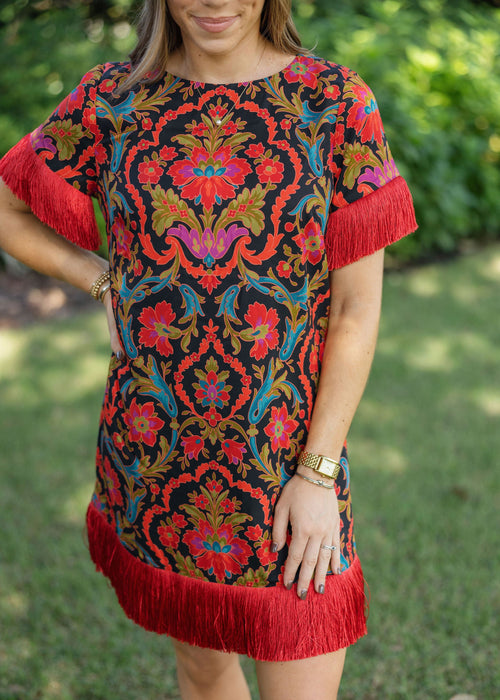 A woman wearing a short-sleeve black dress with a vibrant red, orange, and blue floral damask pattern and red fringe trim on the sleeves and hem, photographed outdoors on a grassy lawn.