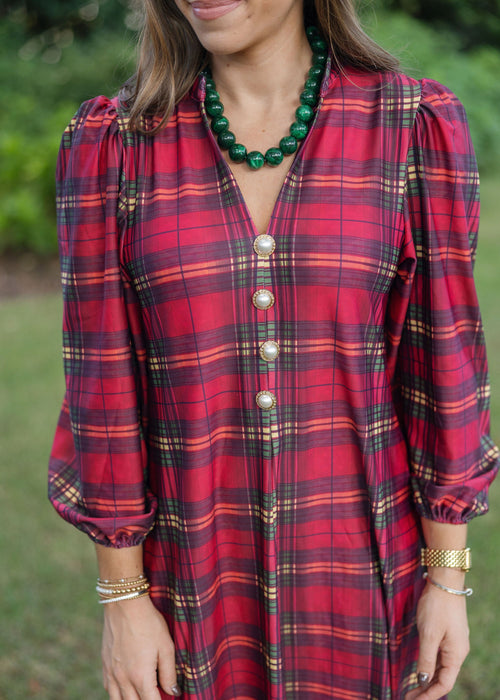 A woman wearing a red and black plaid long-sleeve dress with gold buttons, styled with a green beaded necklace and gold bracelets, set against a soft-focus green outdoor background.