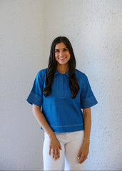 Woman wearing a blue top and white pants against a light gray wall