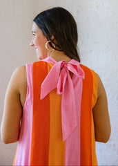 Woman wearing a pink and orange striped dress with a pink bow on the back against a beige wall.