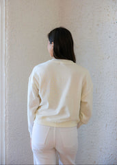 Person wearing a light-colored sweater and pants against a textured white wall