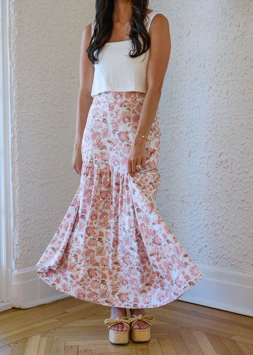 Woman wearing a white top and floral skirt standing against a white wall.