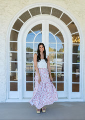 Woman in a white top and floral skirt standing in front of a large arched window.