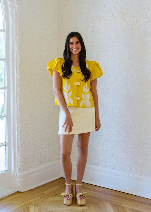 Woman in a yellow floral blouse and white skirt standing in a room with a window.