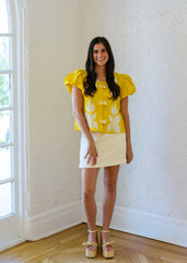 Woman in a yellow floral blouse and white skirt standing in a room with a window.