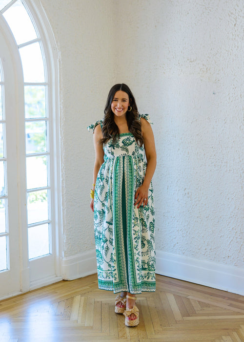 Woman in a green and white patterned dress standing in a room with white walls and wooden floor.