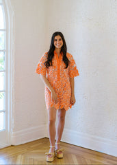 Woman wearing an orange dress with a floral pattern in a room with white walls and wooden flooring.