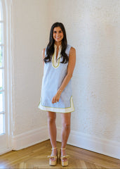 Woman wearing a light blue dress with yellow trim standing in a room with white walls and wooden flooring.