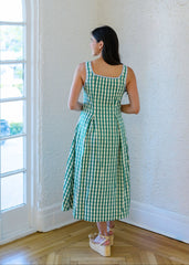Woman in a green checkered dress standing in a room with white walls and wooden floor.