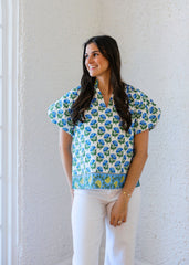 Woman wearing a floral blouse and white pants standing against a white wall.