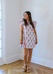 Woman wearing a pink dress with a pineapple pattern in a room with white walls and wooden flooring.