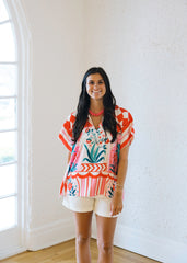 Woman wearing a colorful patterned top and white shorts standing in a room with a white wall and window.