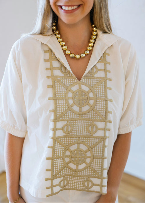 Woman wearing a cream-colored blouse with intricate gold patterns and a gold necklace.