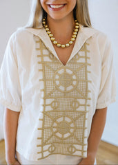 Woman wearing a cream-colored blouse with intricate gold patterns and a gold necklace.