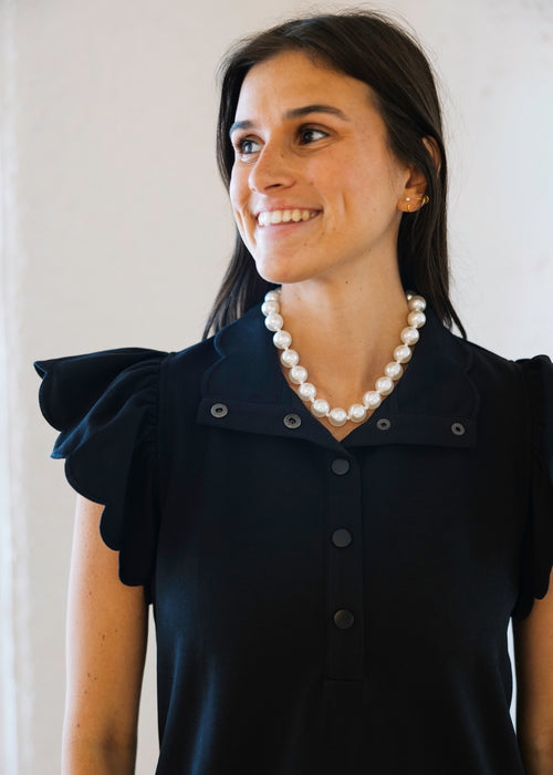 Woman wearing a black dress with ruffled shoulders and a pearl necklace against a white background