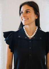 Woman wearing a black dress with ruffled shoulders and a pearl necklace against a white background