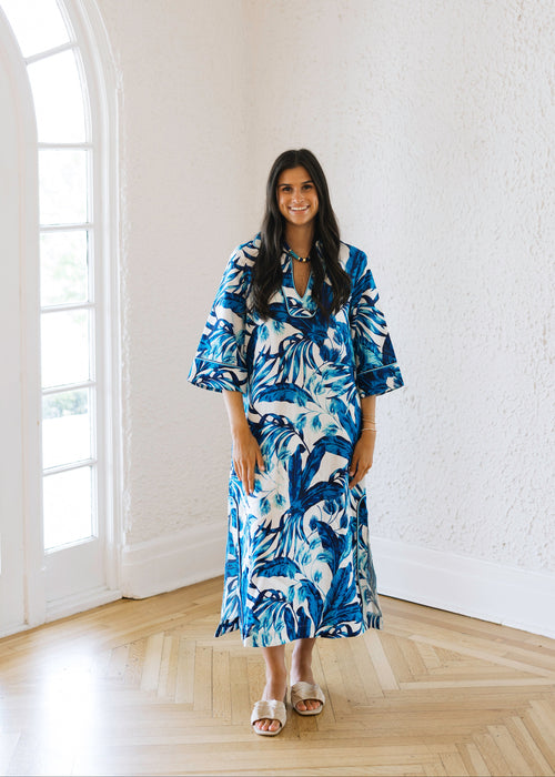 Woman in a blue and white patterned dress standing in a bright room with large windows.