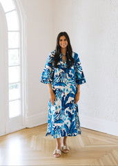 Woman in a blue and white patterned dress standing in a bright room with large windows.