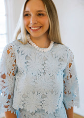 Woman wearing a light blue lace dress with a white pearl necklace indoors.