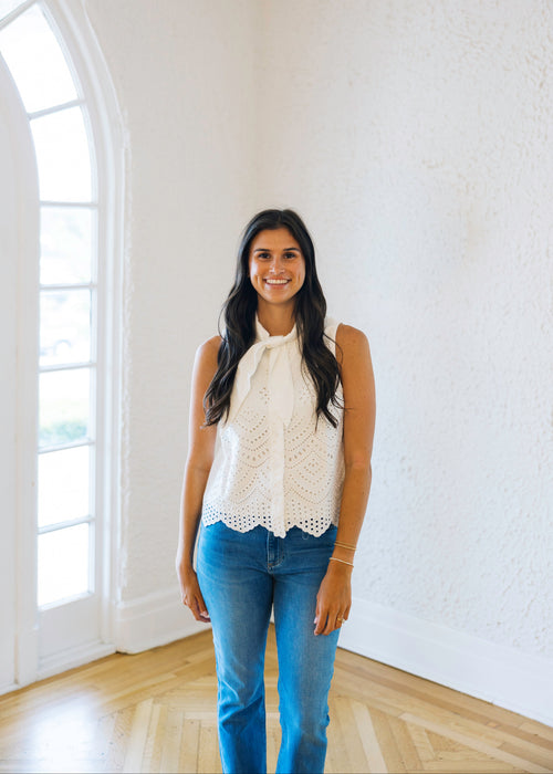 Woman wearing a white lace top and blue jeans standing in a bright room with large windows.