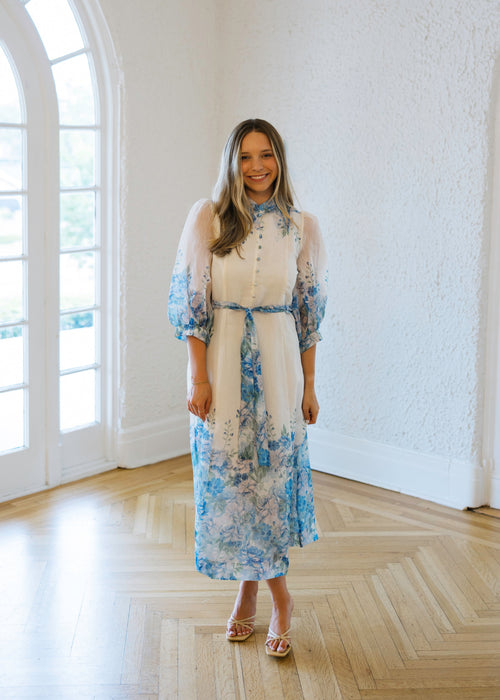 Woman in a floral dress standing in a bright room with large windows.
