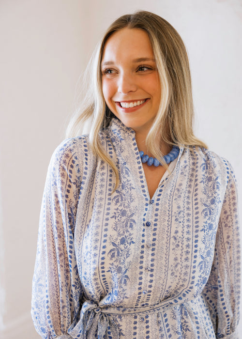 Woman wearing a blue and white patterned dress with a necklace against a plain background