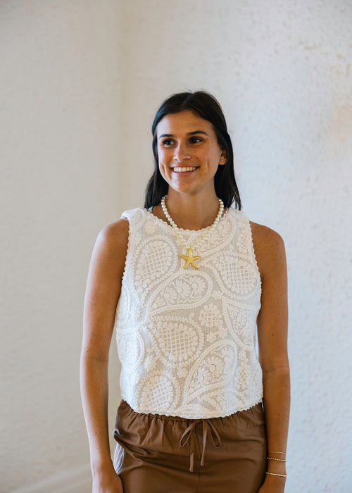 Woman wearing a white sleeveless top with a star design and brown pants against a plain background
