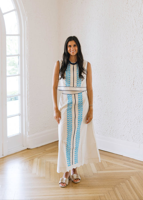 Woman in a white dress with blue patterns standing in a bright room with large windows.