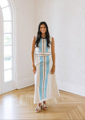 Woman in a white dress with blue patterns standing in a bright room with large windows.