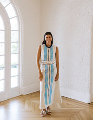 Woman in a white dress with blue patterns standing in a room with large windows.
