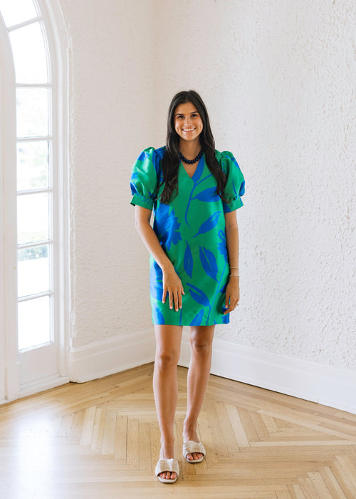 Woman in a blue and green dress standing in a bright room with large windows.