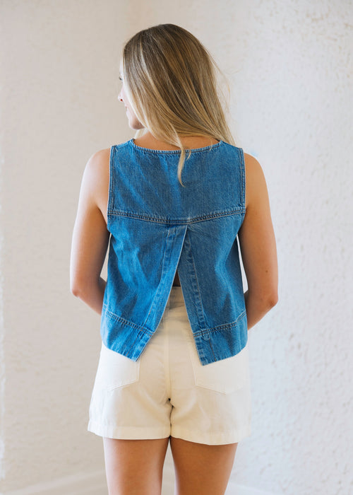 Woman wearing a blue denim vest over a beige top and shorts in a room with white walls and wooden floor.