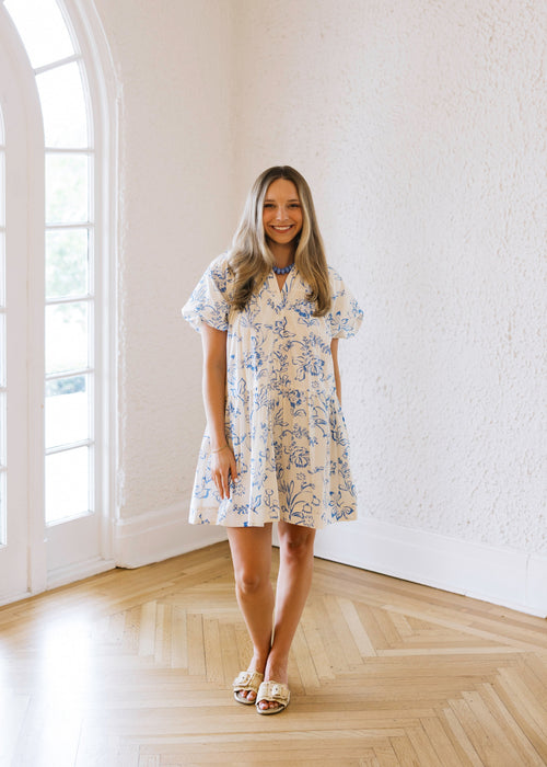 Woman in a floral dress standing in a bright room with large windows.