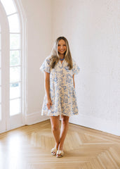 Woman in a floral dress standing in a bright room with large windows.