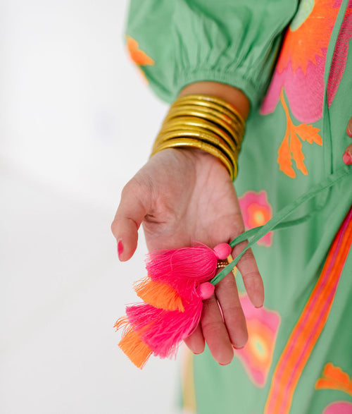 Person wearing a green dress with floral patterns, holding a pink and orange tassel against a white background.